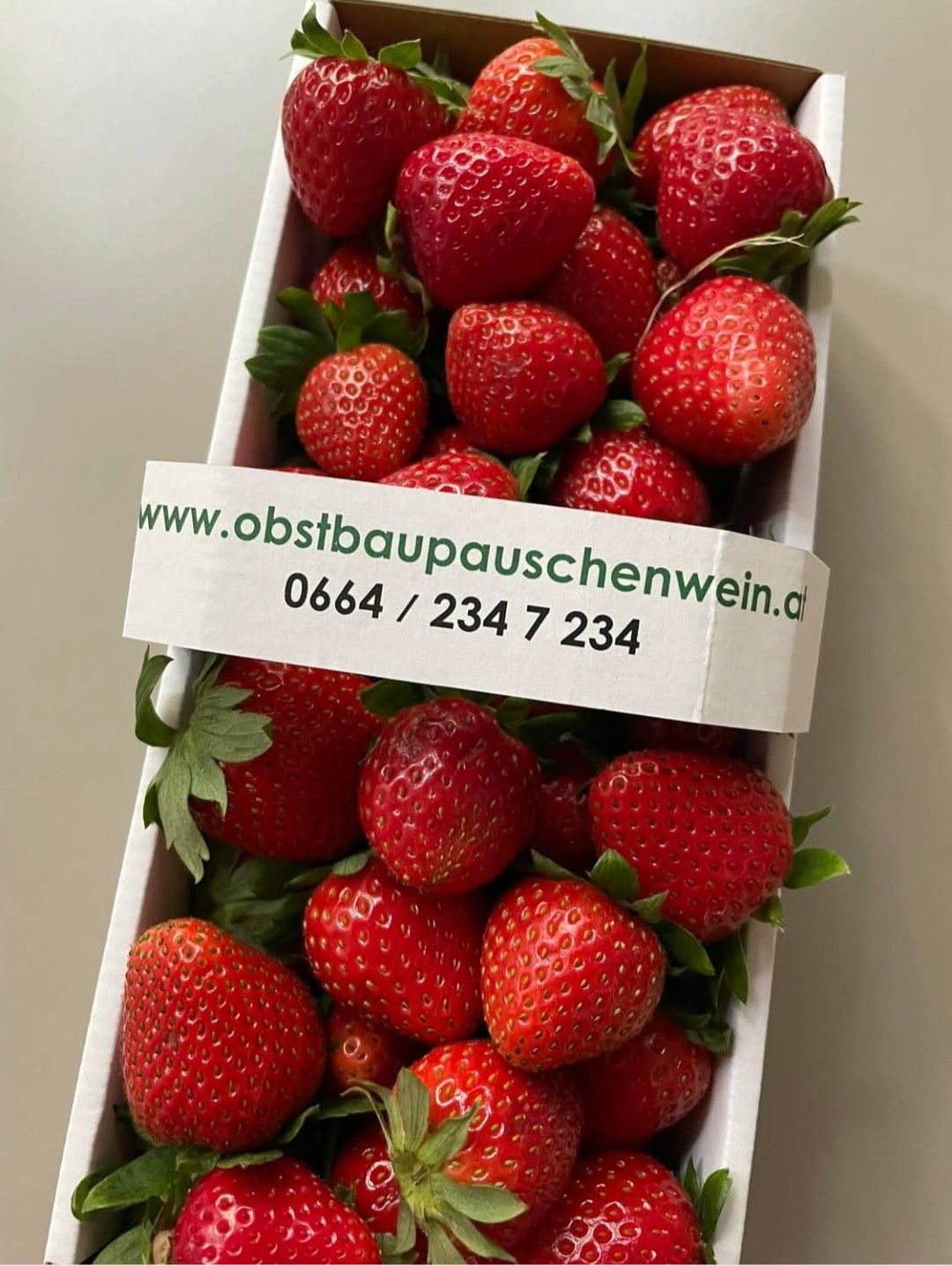 Ein weißer Kasten gefüllt mit roten Erdbeeren. Ein weißes Etikett mit grünem Text darauf sagt www.obstbaupauschenwein.at und die Telefonnummer 0664/234 7 234.