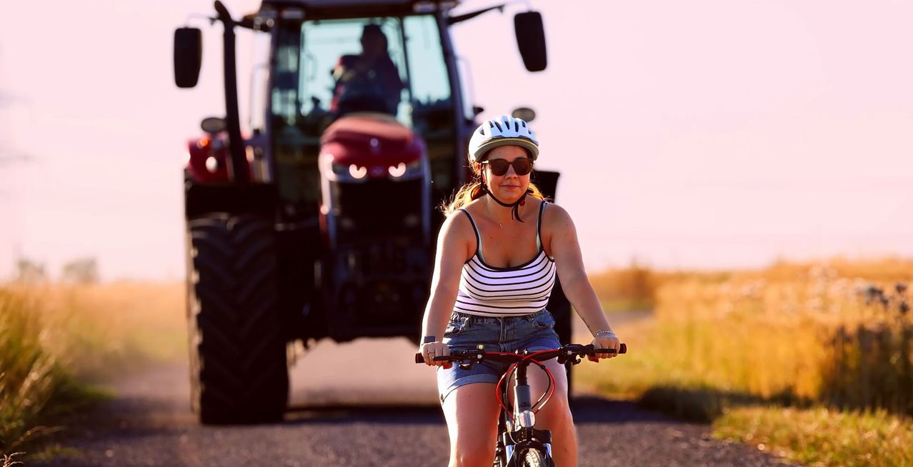 Eine Frau fährt auf einer Landstraße mit einem Traktor im Hintergrund ein Fahrrad. Sie trägt einen Helm und Sonnenbrille.