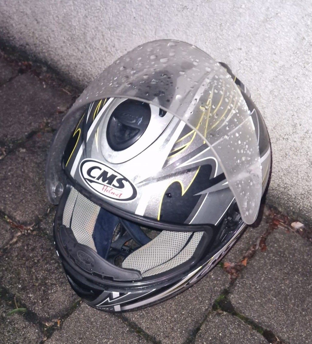 Ein silberner und schwarzer Motorradhelm mit einer transparenten Regenabdeckung, auf einem gepflasterten Gehweg liegend.