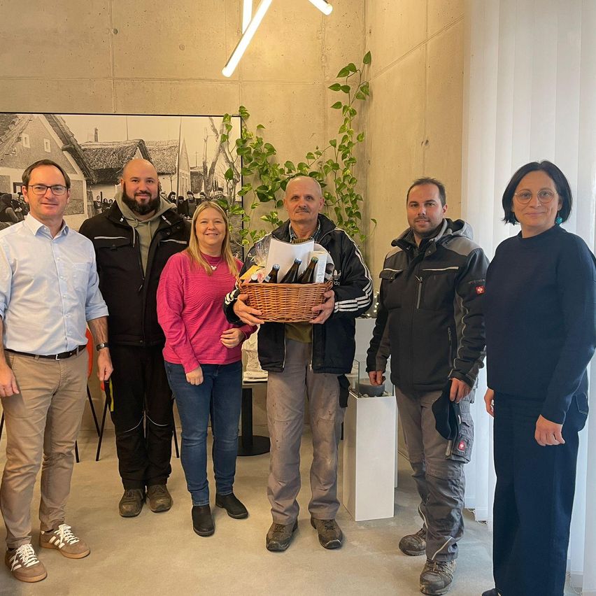 Sechs Menschen, vier Männer und zwei Frauen, posieren für ein Foto. Eine Frau hält einen Korb. Dahinter befindet sich eine Wand mit einem Foto und einer Pflanze.