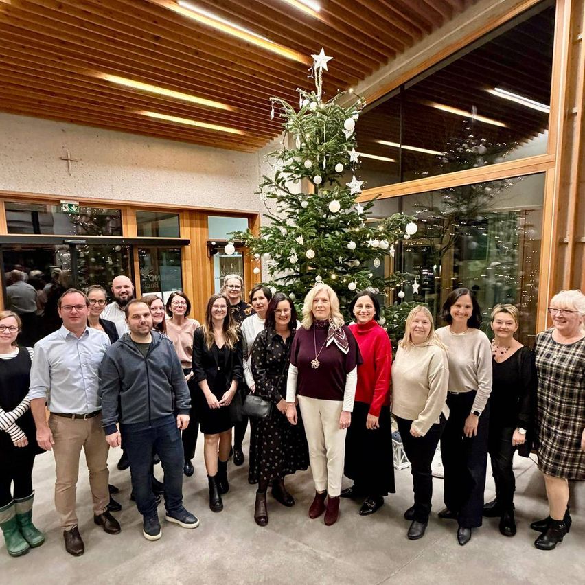 Eine Gruppe von Menschen steht vor einem geschmückten Weihnachtsbaum und posiert für ein Foto. Der Raum hat Holzwände und große Glasfenster.