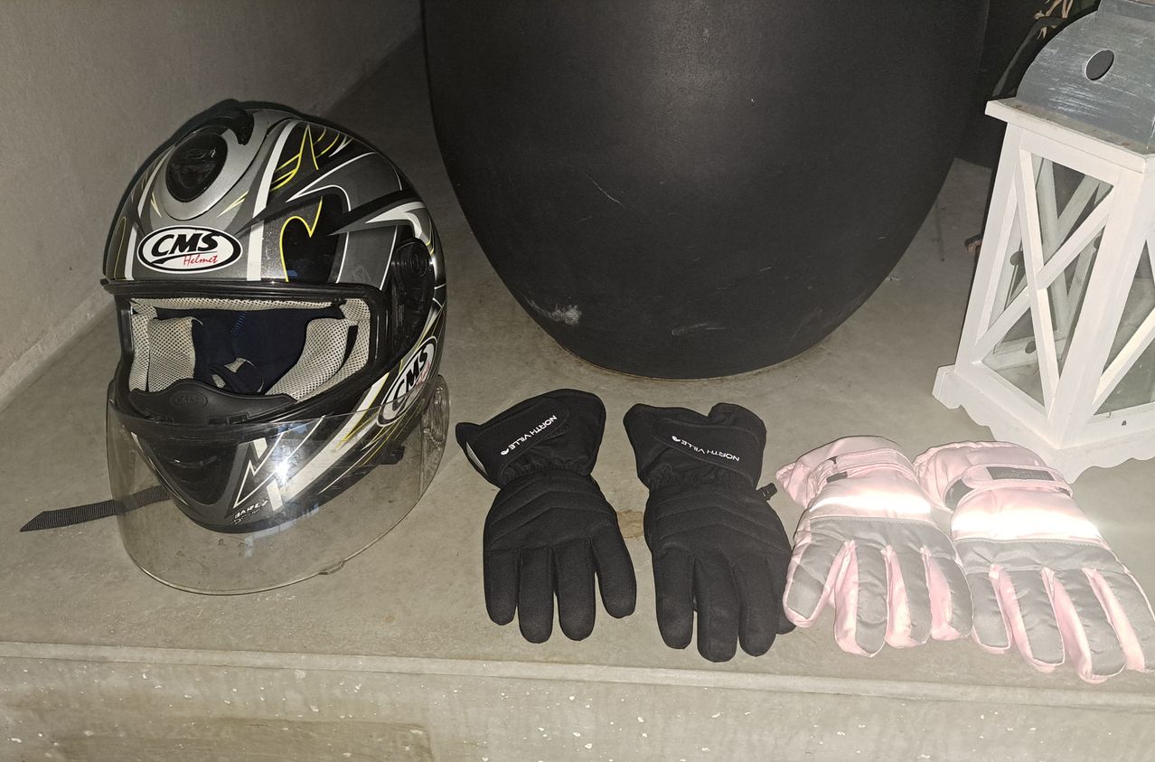 Ein schwarzer Motorradhelm, zwei schwarze Handschuhe und ein rosa-grauer Handschuh liegen auf einer Betonoberfläche. Ein großer schwarzer Gegenstand befindet sich hinter dem Helm.