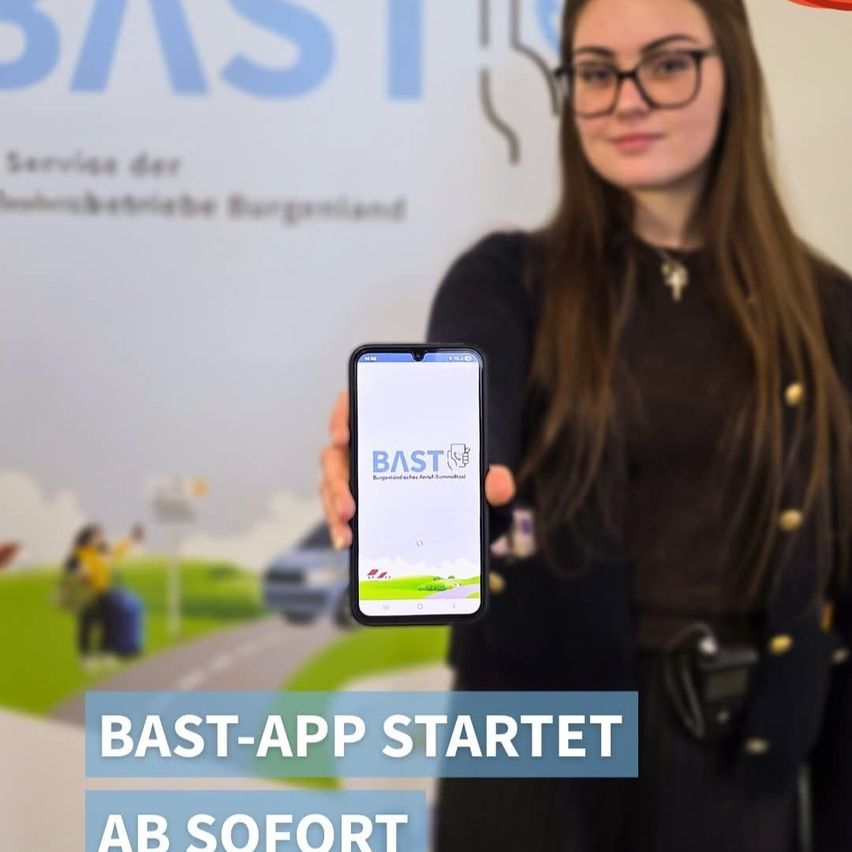 Eine Frau mit Brille hält ein Smartphone mit der BAST-App. Das Logo der App und ein Schild mit 'BAST-APP STARTET AB SOFORT' sind sichtbar. Ein unscharfes Bild von einem Auto und einem Fußgänger ist im Hintergrund.