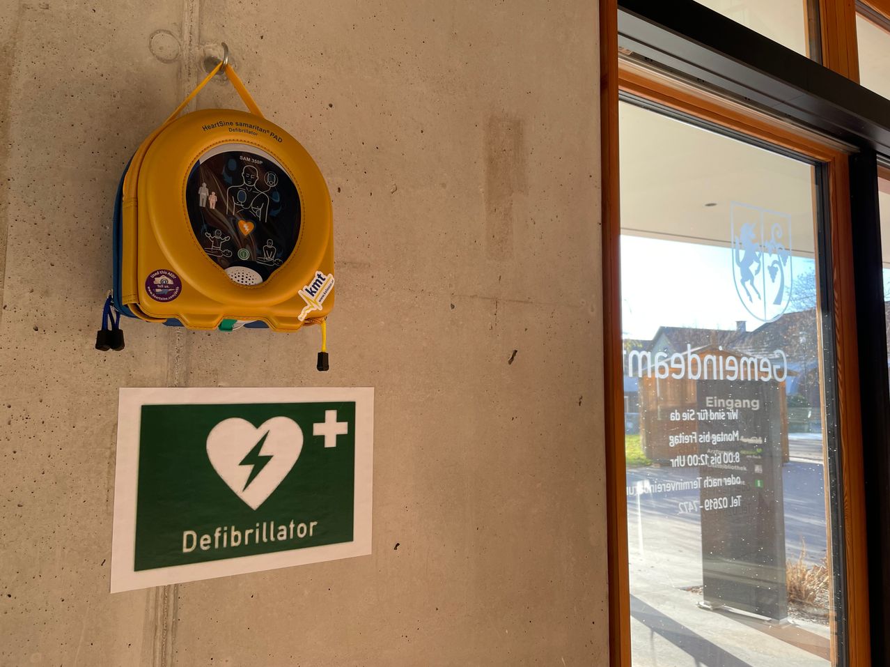 Ein Defibrillator ist an einer Wand montiert, mit einem Schild darunter, das seine Anwesenheit anzeigt. Der Defibrillator hat ein gelbes Gehäuse und einen blauen Griff. Das Schild ist grün mit einem weißen Herz und Blitzsymbol.