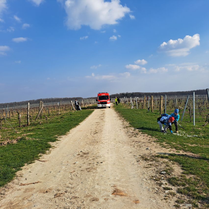 A red emergency vehicle drives down a dirt road. People are standing by a fence and one is picking up something.