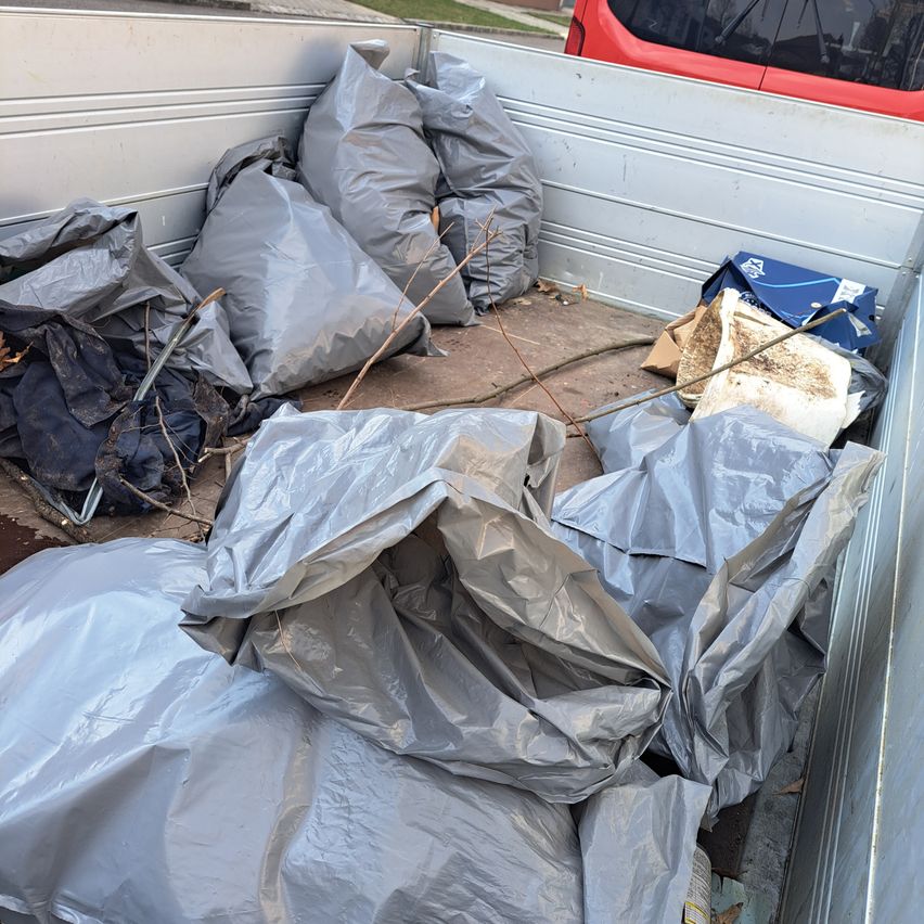 A truck filled with gray plastic bags, some containing branches, and other debris. A red car is parked nearby.