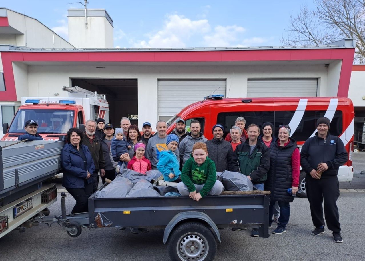 A group of people are gathered around a trailer filled with bags of trash, posing for a photo in front of a fire station.