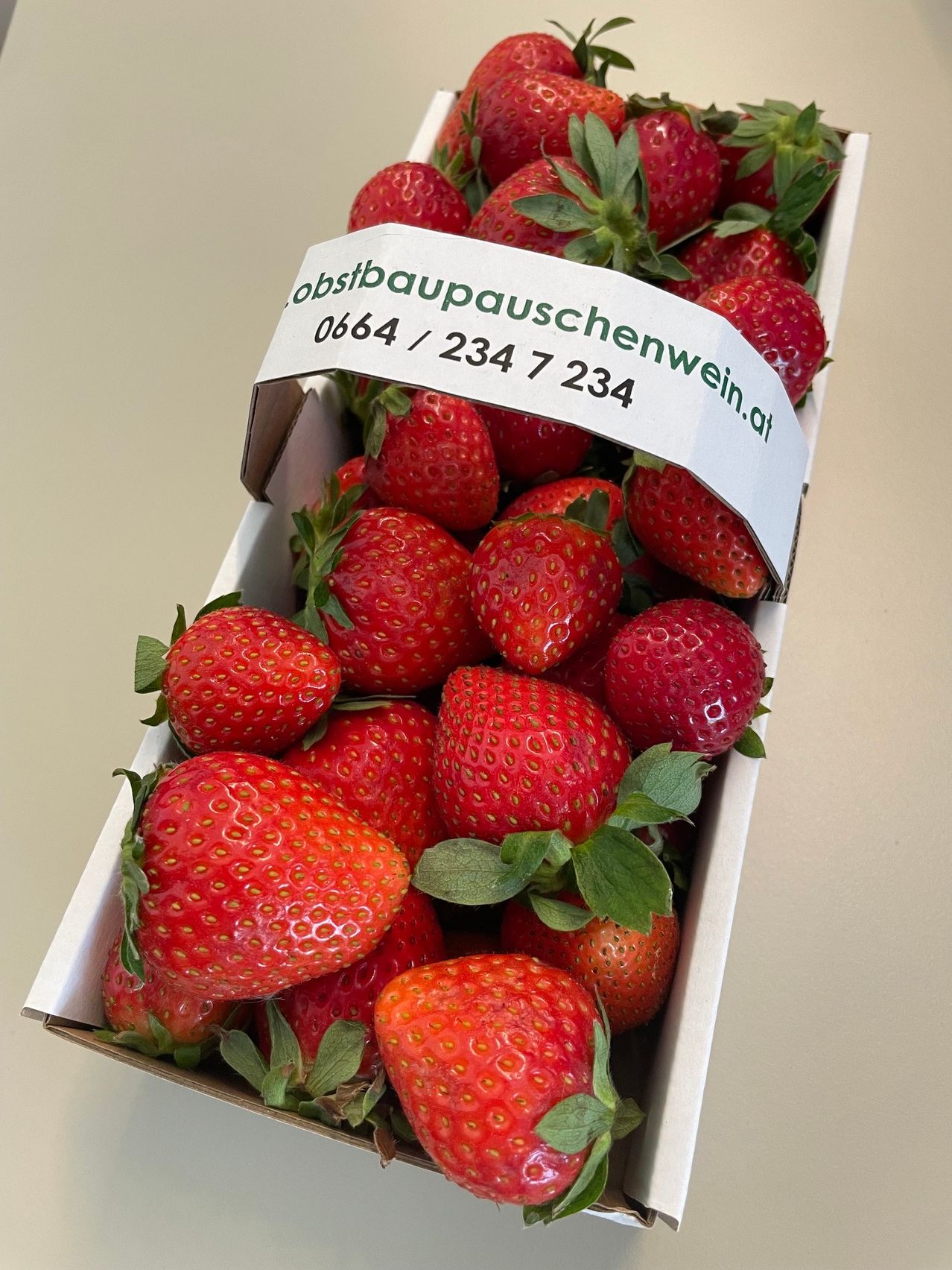 Eine Schachtel roter Erdbeeren mit grünen Blättern, beschriftet mit einer Telefonnummer.