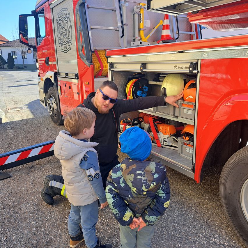 A man is showing two kids the equipment in a fire truck. The truck is parked on a gravel road.