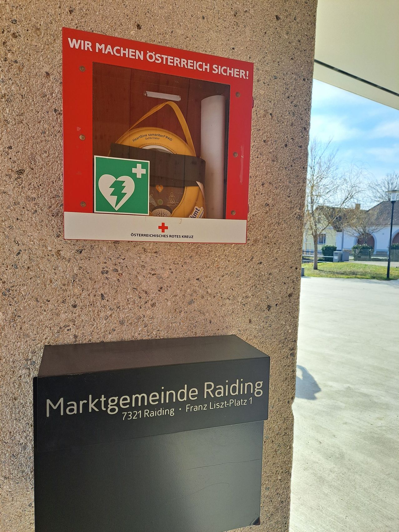 Eine rote und weiße Box, die an einer Wand montiert ist, enthält einen gelben Rettungsring. Darunter steht ein schwarzes Schild mit der Aufschrift Marktgemeinde Raiding 7321 Raiding. Draußen ist ein Rasenbereich mit einem Gebäude und einem Baum zu sehen.