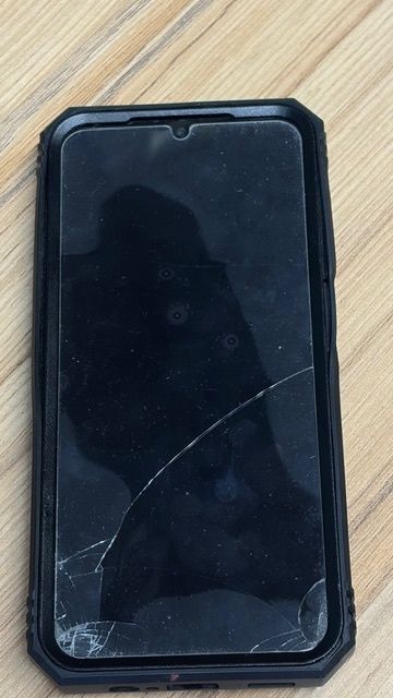 Die Rückseite einer schwarzen Smartphone-Hülle mit einem gesprungenen Bildschirm, auf einer Holzoberfläche platziert.