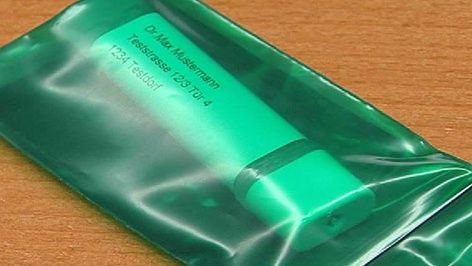Eine grüne Tube mit der Aufschrift Dr. Max Mustermann Testsubstanz 123b, 704 befindet sich in einer durchsichtigen Plastiktüte auf einem Holztisch.