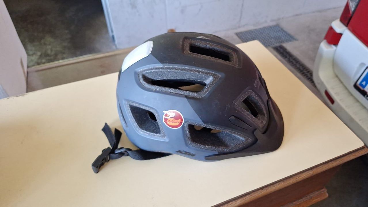 A black bicycle helmet with a red logo is displayed on a table. It has multiple air vents and a strap.