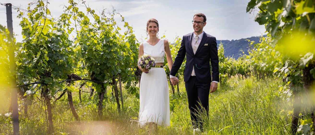 Ein frisch verheiratetes Paar geht lächelnd Hand in Hand durch einen Weinberg. Die Frau trägt ein weißes Kleid, und der Mann ist in einem Anzug.