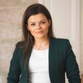 Eine lächelnde Frau mit langen braunen Haaren steht vor einer weißen Wand. Sie trägt ein weißes Shirt, einen grünen Blazer und eine Halskette.