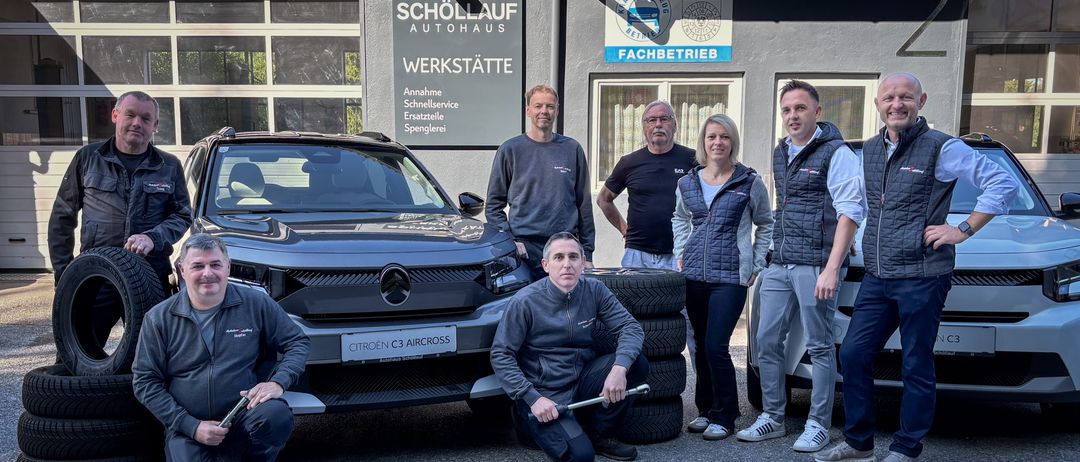 Eine Gruppe von Männern und Frauen steht vor einem Gebäude mit einem Schild, das 'Schöllauf Werkstatte' und ein Citroen C3 Aircross Auto zeigt. Die Männer halten Werkzeuge und einer sitzt auf dem Boden.
