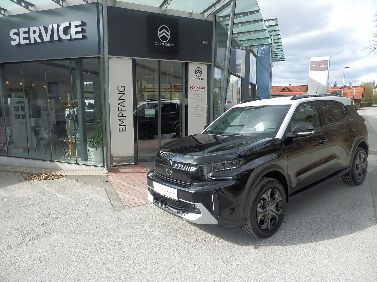 Ein schwarz-weißer SUV steht vor einem Citroen-Autohaus, mit einer Glaswand und einem Topfpflanzen im Inneren sichtbar.