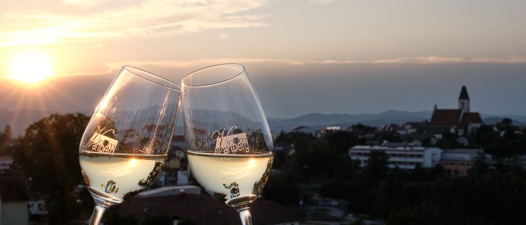 Zwei Gläser Wein auf einem Hügel mit einem Sonnenuntergangshimmel und Stadtkulisse im Hintergrund. Der Wein spiegelt die Landschaft und Berge wider.