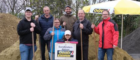 Eine Gruppe von Menschen in Jacken und Brillen mit Schaufeln posiert für ein Foto mit einem kleinen Mädchen in der Mitte, das eine Mütze trägt. Hinter ihnen steht ein Schild mit der Aufschrift 'Sandkasten-Aktion'.