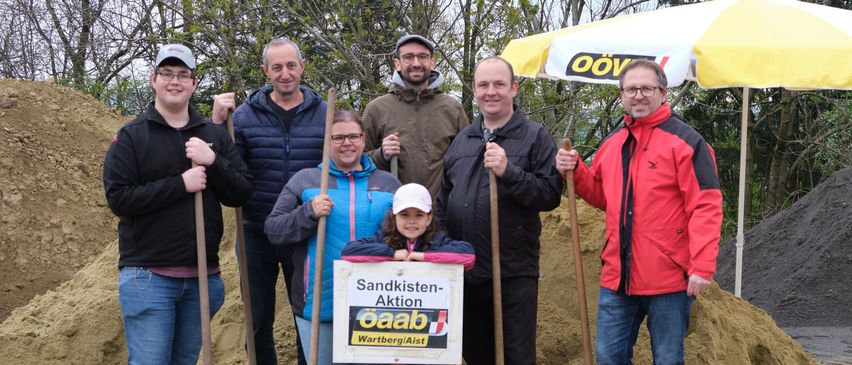 Eine Gruppe von Menschen in Jacken und Brillen mit Schaufeln posiert für ein Foto mit einem kleinen Mädchen in der Mitte, das eine Mütze trägt. Hinter ihnen steht ein Schild mit der Aufschrift 'Sandkasten-Aktion'.