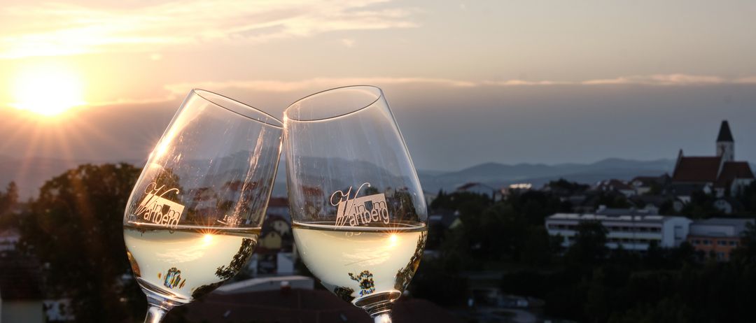 Zwei Weingläser mit weißem Wein, beschriftet mit 'Wanberg', werden vor einem Hintergrund von untergehender Sonne und Bergen gehalten.