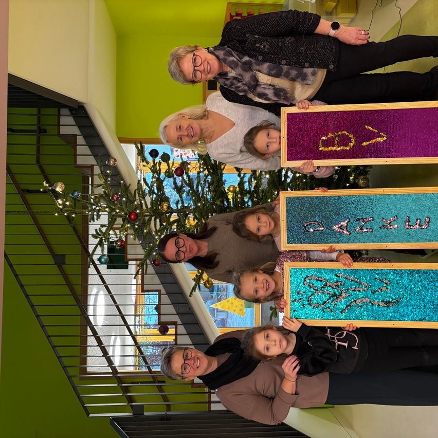 Eine Gruppe von Erwachsenen und Kindern steht zusammen vor einem geschmückten Weihnachtsbaum. Jede Person hält ein farbenfrohes Board mit Pailletten. Der Hintergrund zeigt eine Inneneinrichtung mit einer grünen Wand und einer Treppe.