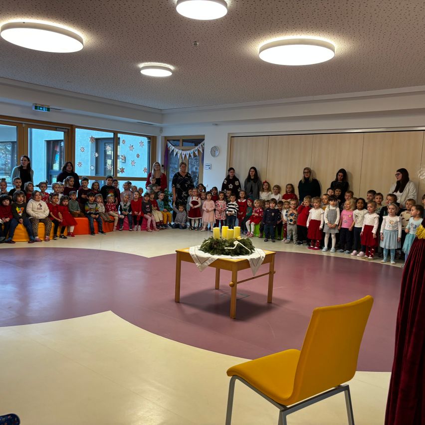 In einem großen Raum sind viele Kinder versammelt, einige sitzen auf einer Bank, andere stehen. Ein Tisch mit Kerzen und Grün steht in der Mitte. Erwachsene sind anwesend, und es herrscht eine festliche Atmosphäre.