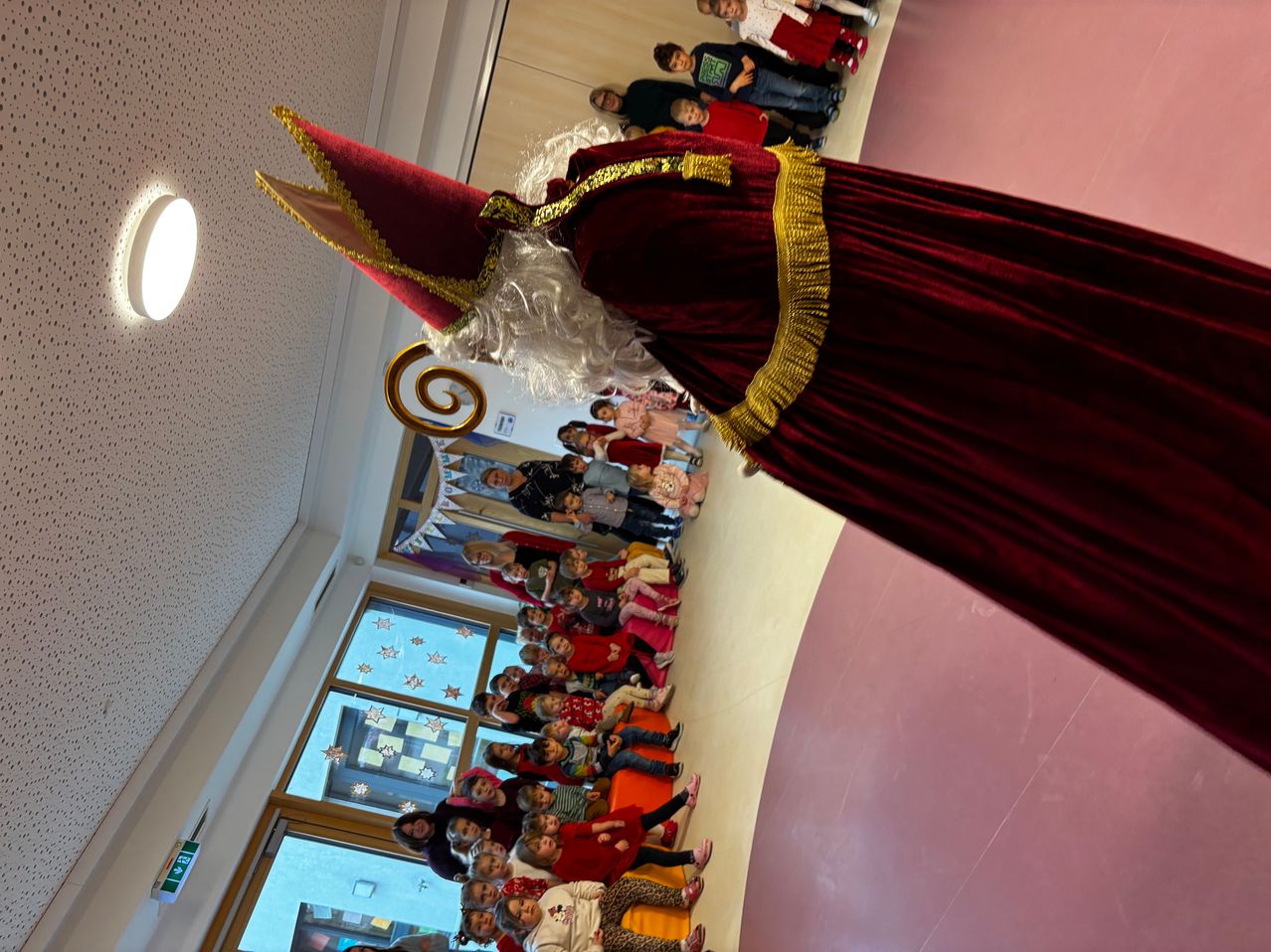 Eine Person in einem roten Kostüm mit einem spitzen Hut steht vor einer Gruppe von Kindern, mit einer gemusterten Wand und Glasfenstern im Hintergrund.