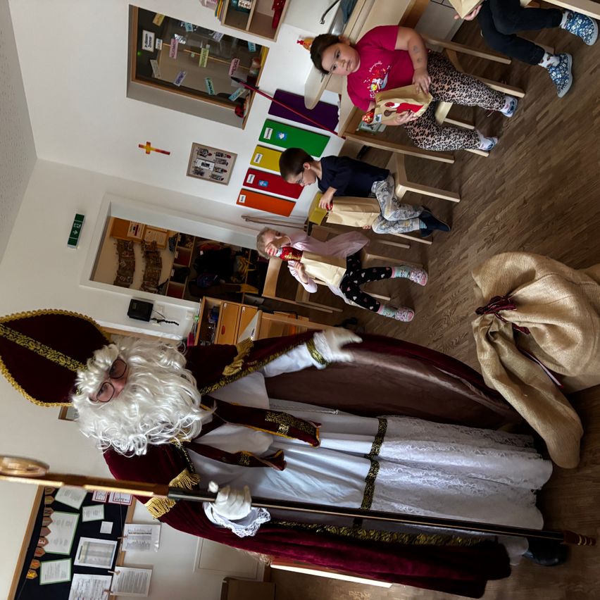 Eine Person in Santa-Claus-Verkleidung steht in einem Klassenzimmer mit Kindern, die an Schreibtischen sitzen und Geschenktüten betrachten. Bunte Poster und Bulletin-Boards hängen an den Wänden.