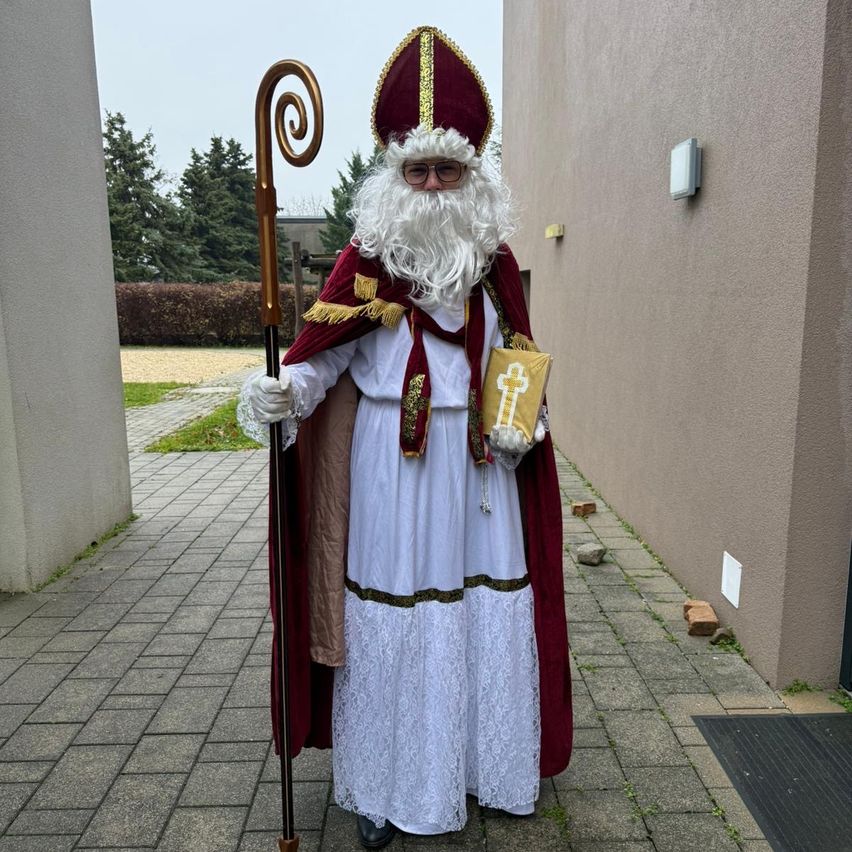 Eine Person verkleidet als Bischof mit einem Kreuz auf der Brust und einem Buch in der Hand, steht auf einem gepflasterten Boden.
