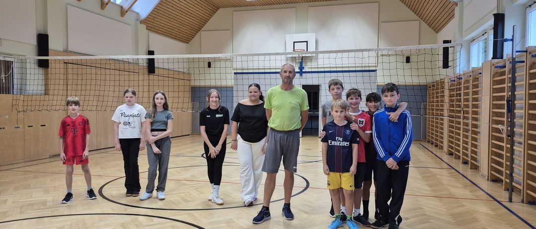 Eine Gruppe von Personen, darunter ein Mann, zwei Frauen und mehrere Kinder, stehen gemeinsam in einer Turnhalle und lächeln für ein Foto.