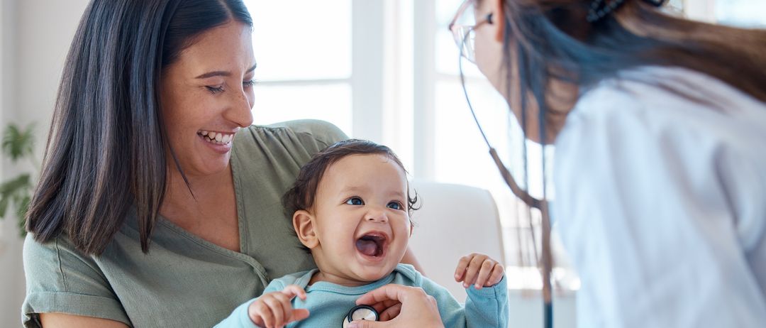 Ein Baby lacht, während eine Ärztin es mit einem Stethoskop untersucht, gehalten von einer lächelnden Mutter.