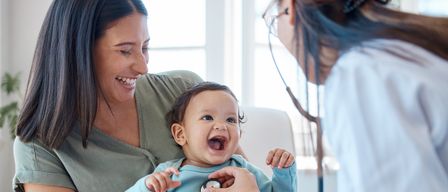 Ein Baby lacht, während eine Ärztin es mit einem Stethoskop untersucht, gehalten von einer lächelnden Mutter.