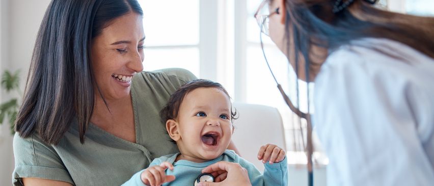 Ein Baby lacht, während eine Ärztin es mit einem Stethoskop untersucht, gehalten von einer lächelnden Mutter.