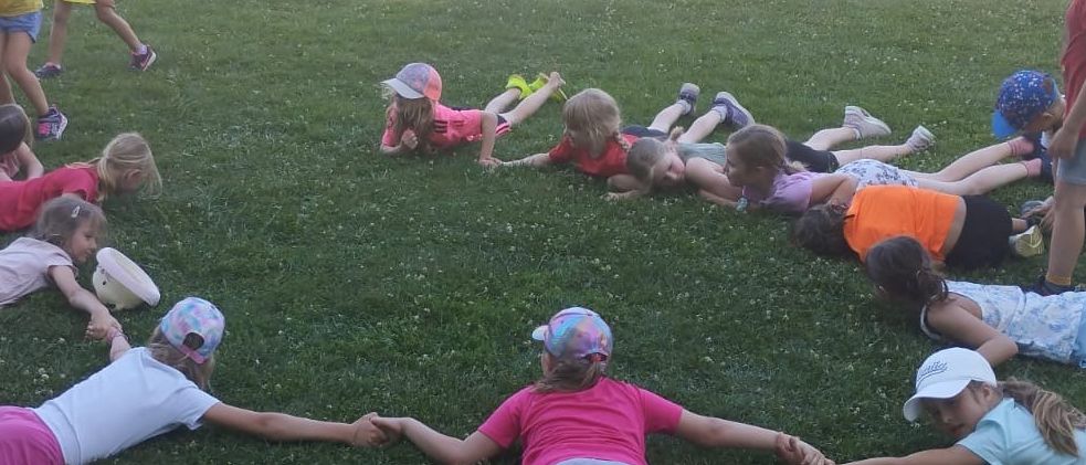 Kinder liegen im Kreis auf dem Rasen. Einige tragen Mützen. Dahinter befinden sich Gebäude und Bäume. Das Bild ist vom 12.06.2021.