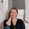 Eine Frau sitzt vor einer Tür auf dem Boden, lächelt und trägt ein schwarzes Shirt und Jeans.