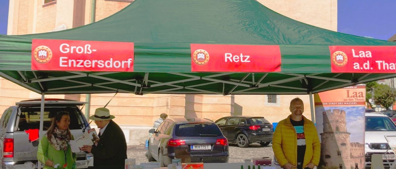 Ein Stand mit einem grünen Zelt, auf dem die Worte 'Groß-Enzersdorf' und 'Retz' stehen. Zwei Personen stehen hinter einem Tisch mit Flaschen und anderen Artikeln. Ein Gebäude steht hinter ihnen.