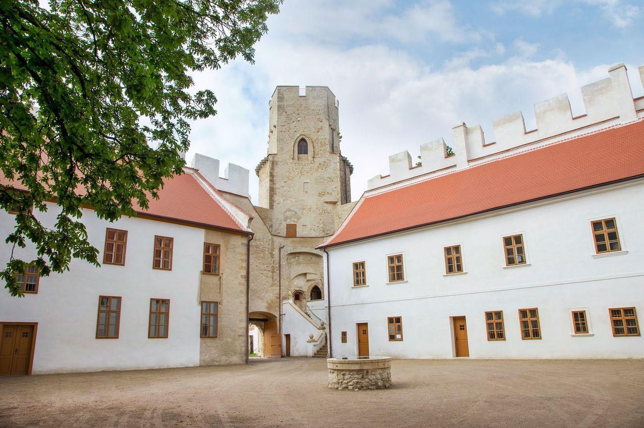 Eine große mittelalterliche Burg mit roten Dächern und einem Steinturm in der Mitte. Ein großer Innenhof vor der Burg mit einem Steinbrunnen in der Mitte.