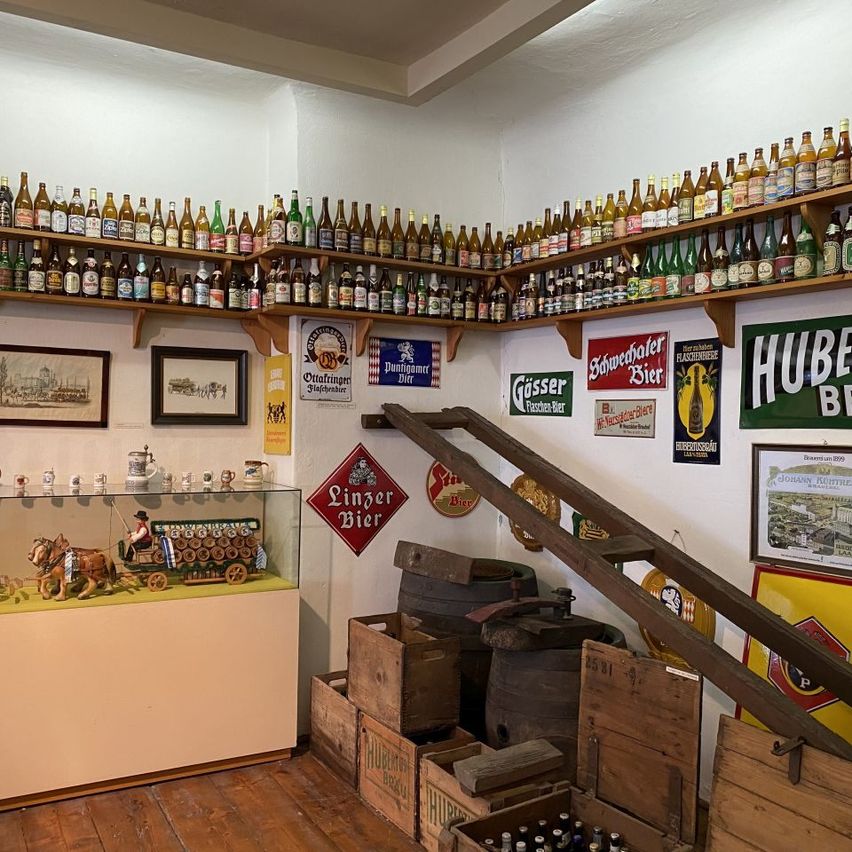 Ein Raum mit Holzregalen, die mit vielen Bierflaschen gefüllt sind, einem Glaskasten mit einem Pferdefuhrwerkmodell und Holzkisten.