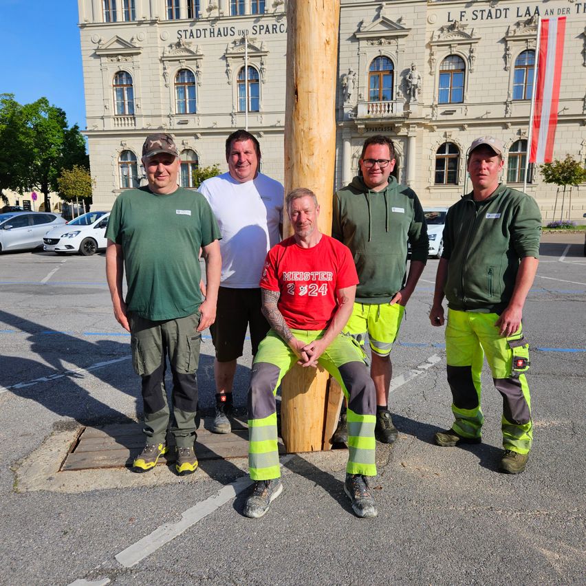 Fünf Männer in grüner Arbeitskleidung posieren für ein Foto vor einem großen Gebäude mit mehreren Fenstern und einer Flagge.