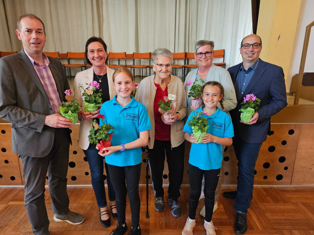 Eine Gruppe von Menschen, darunter zwei junge Mädchen, steht in einem Raum und hält Topfpflanzen. Hinter ihnen stehen drei ältere Frauen, eine mit einem Gehstock, vor einem weißen Vorhang.