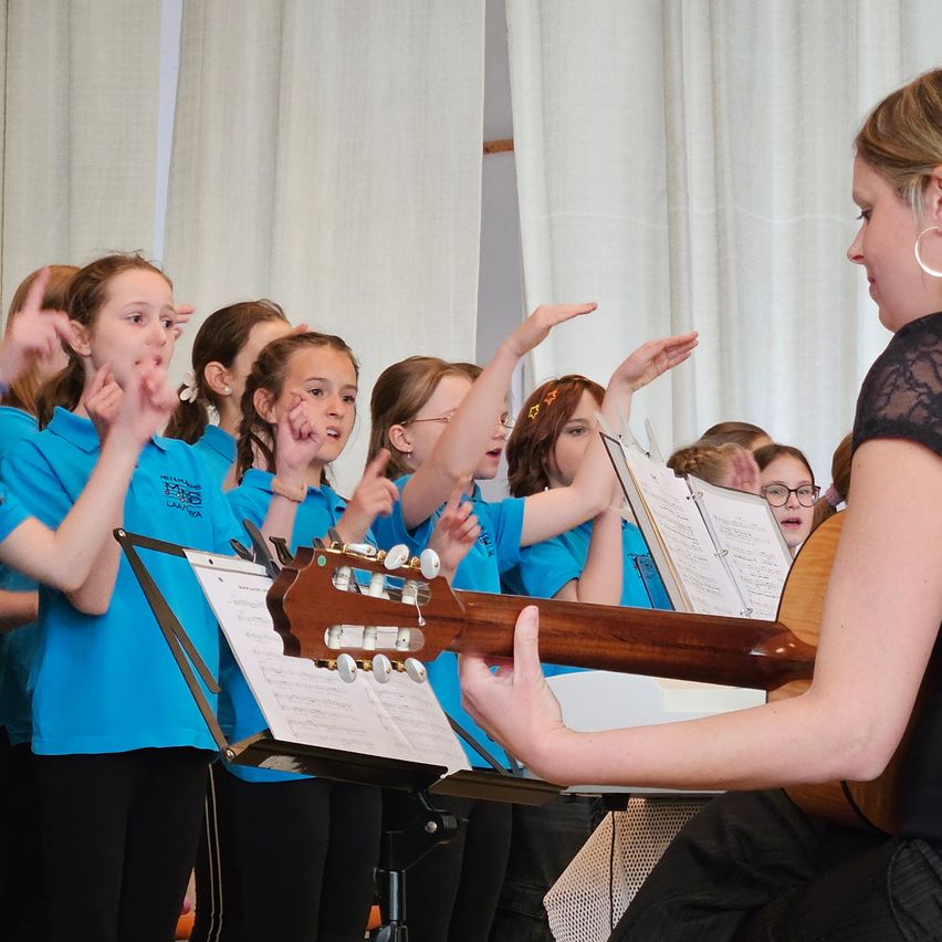 Eine Gruppe junger Mädchen in blauen Shirts singt zusammen mit einer erwachsenen Frau, die Gitarre spielt. Sie stehen vor einem weißen Vorhang.