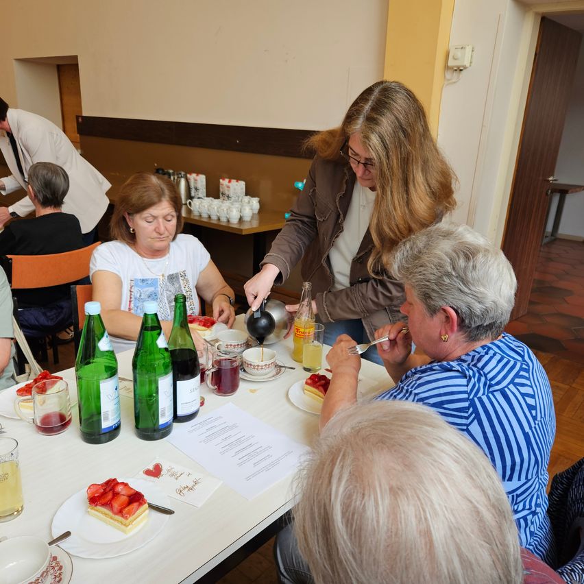 Eine Gruppe älterer Frauen genießt eine Mahlzeit an einem Tisch mit Desserts, Wein und Getränken. Eine Frau schenkt Kaffee ein, während eine andere isst. Ein Mann steht im Hintergrund.