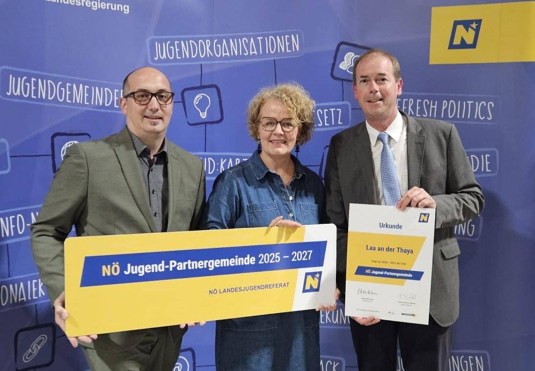 Three people stand together, smiling, in front of a blue backdrop. Two men hold a certificate and a banner. The banner reads 'NO Jugend-Partnergemeinde 2025 - 2027'. The woman wears glasses.