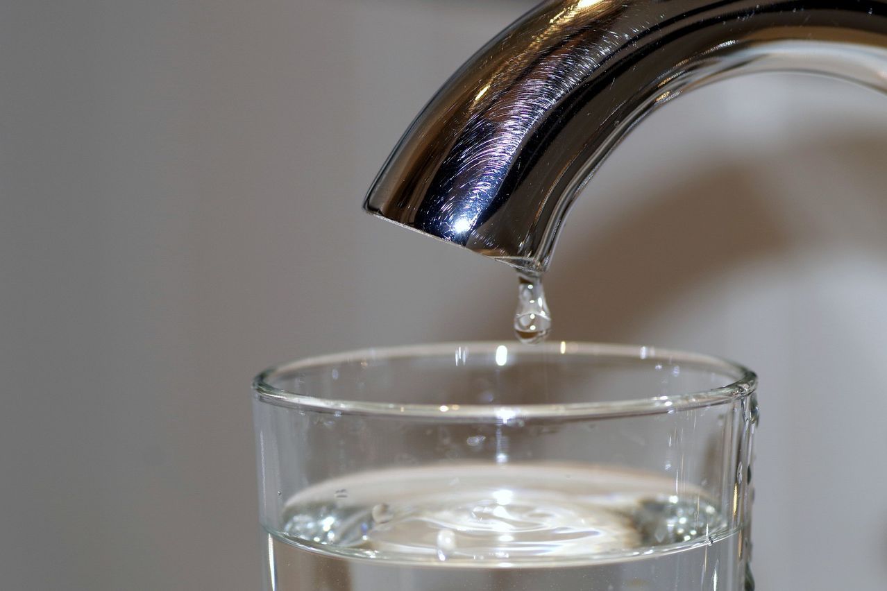 A close-up of a stainless steel faucet dripping water into a clear glass filled with water.