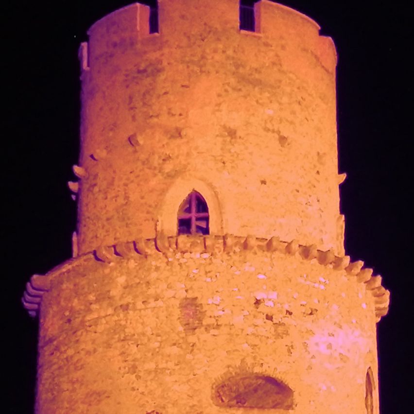 Ein hoher, alter Steinturm, der bei Nacht beleuchtet ist, mit einem Fenster und einer gekrönten Spitze.