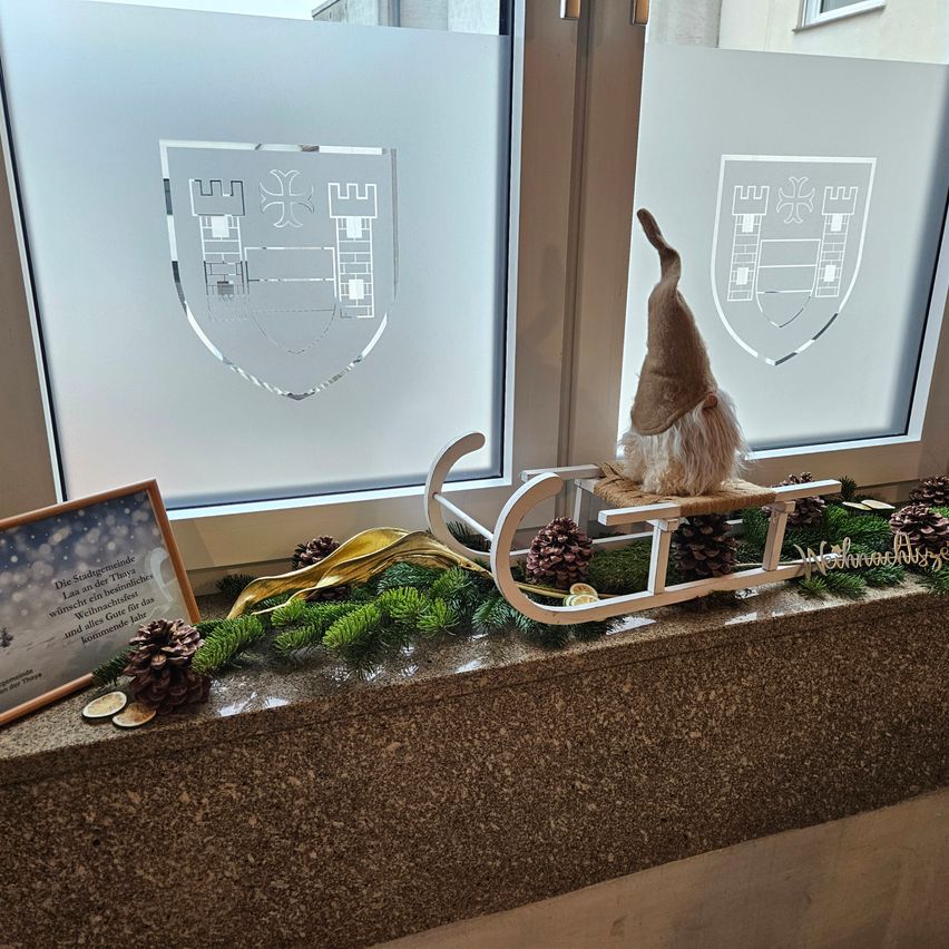 Ein Gnom auf einem Schlitten sitzt auf einer Fensterbank mit Tannenzapfen und Grün, neben einer gerahmten Nachricht. Dahinter sind zwei mattierte Glasscheiben mit einem Wappen zu sehen.