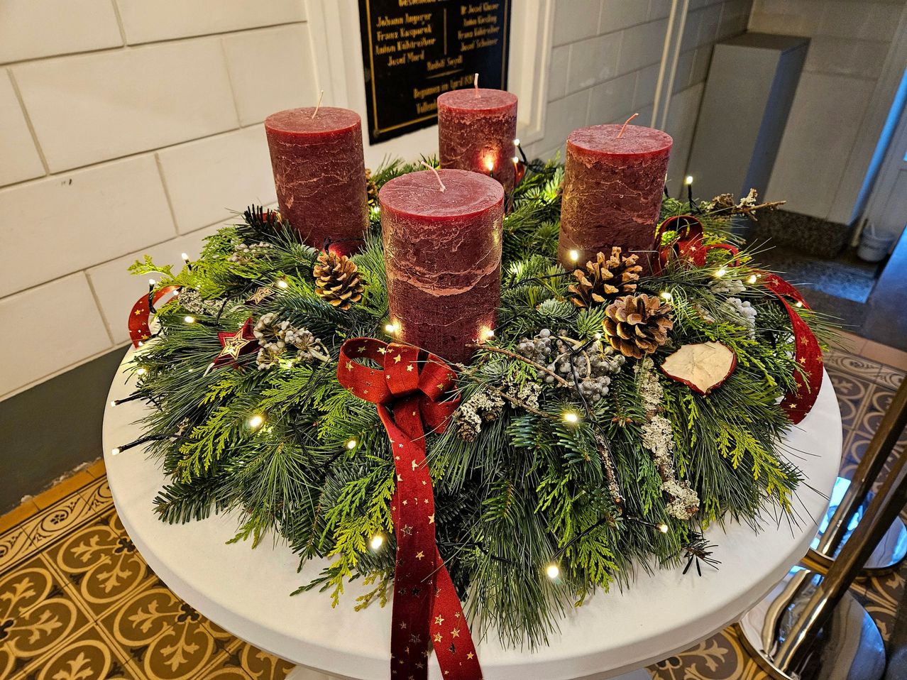 Ein Weihnachtskranz auf einem Tisch mit vier roten Kerzen, einem roten Band und Weihnachtsdekoration. Der Kranz ist mit Tannenzapfen, Lichtern und Grünzeug geschmückt.