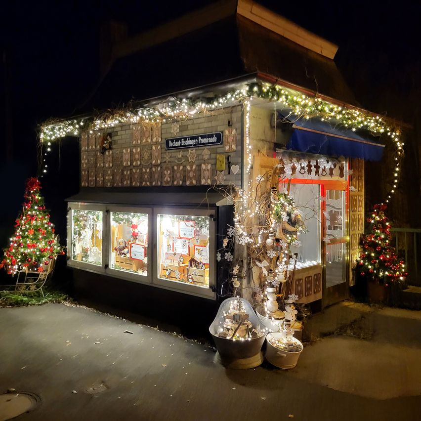 Ein kleines Geschäft mit einem Weihnachtsbaum davor ist mit Lichtern und Ornamenten geschmückt. Das Schild sagt 'Bechhand-Bechlinger-Promenade'. Zwei weitere Weihnachtsbäume sind in der Nähe.