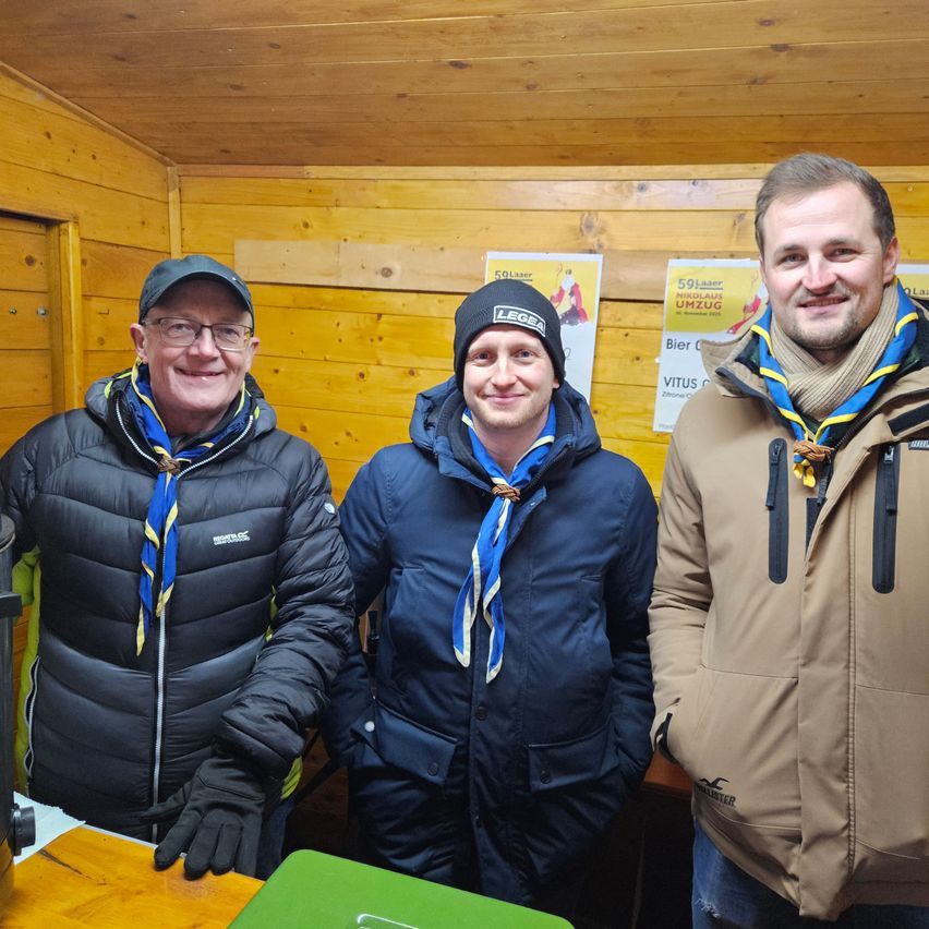 Drei Männer stehen in einer Holzhütte, alle tragen Jacken und Schals. Der Mann auf der linken Seite trägt eine Kappe und eine Brille, der in der Mitte eine schwarze Mütze und der auf der rechten Seite einen Schal. Sie lächeln für ein Foto. Hinter ihnen befinden sich Poster mit Texten.